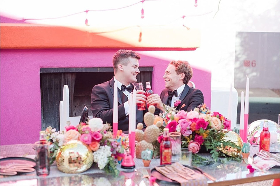 Two grooms cheers to each other sitting at a bright pink tablescape
